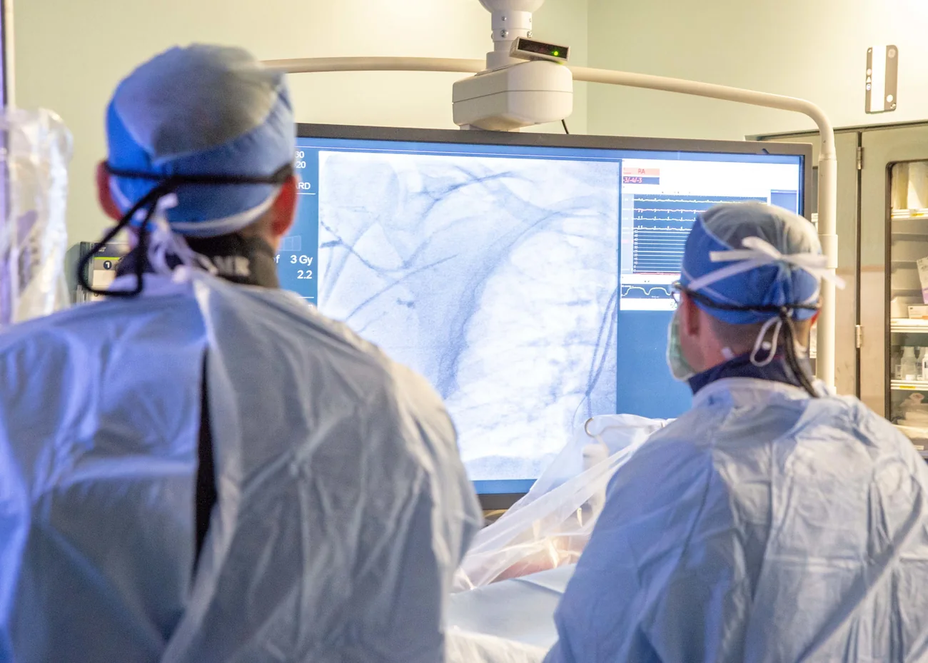 Two clinicians in scrub caps and gowns watching an angiogram on a large screen in a procedure room